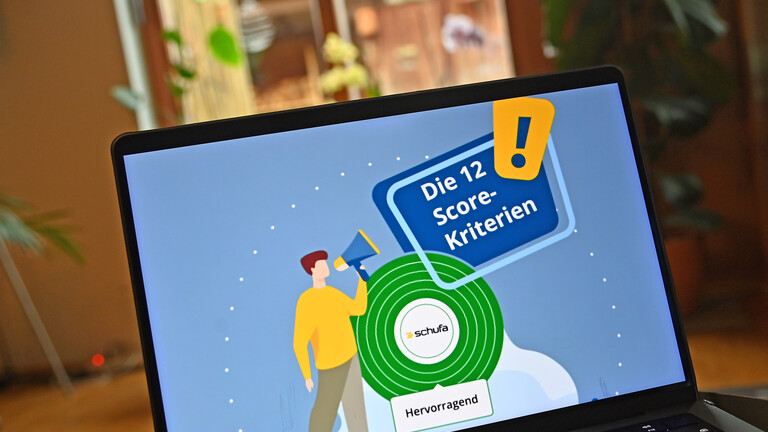 Ein geöffneter Laptop steht in einer Art Wohnzimmer. Im Hintergrund sind Pflanzen zu sehen. Auf dem Bildschirm steht "Die 12 Score-Kriterien". Dieser Schufa-Score schätzt die Kreditwürdigkeit einer Person.