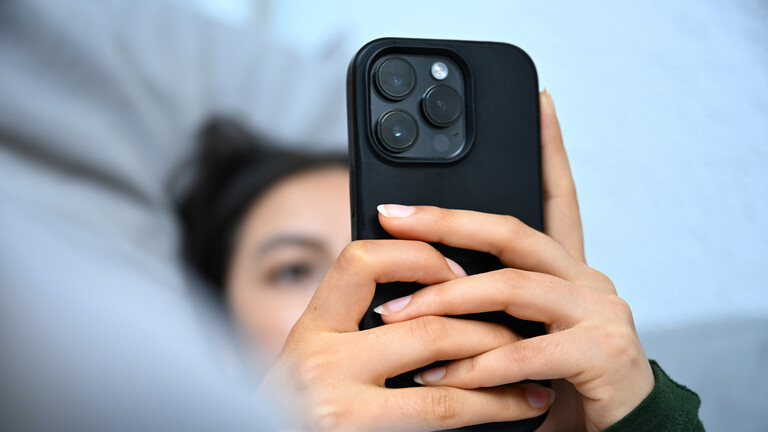 Eine junge Frau liegt mit ihrem Handy auf der Couch und blickt auf das Display.