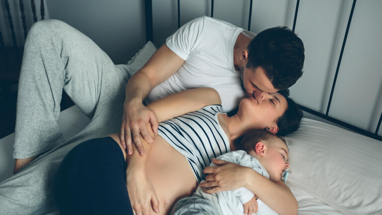 Eine Familie liegt im Bett. Vater, schwangere Mutter plus Kind