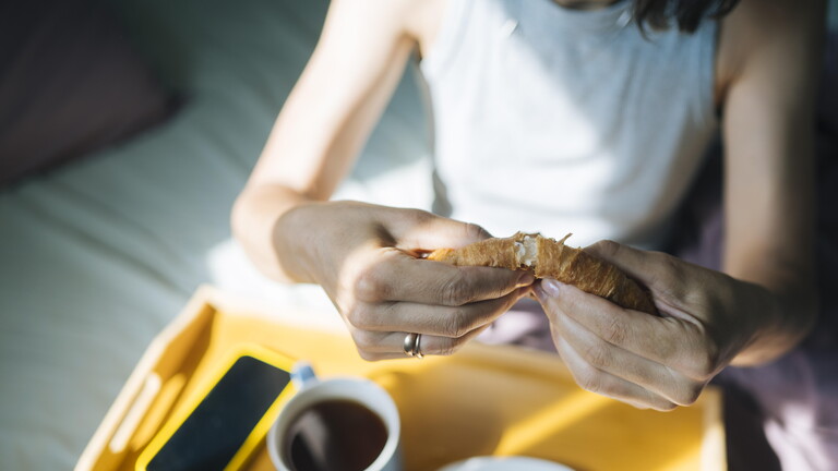 Eine Frau teil ihr Croissant während ihres Frühstücks im Bett. Das Handy liegt parat und eine Tasser Kaffee steht auch herum