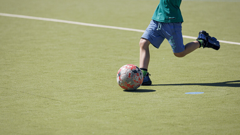 Kind nimmt auf Fußballplatz mit Kunstrasen Anlauf, um Ball zu schießen