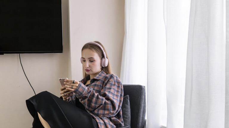 Eine Junge Frau, die Kopfhörer trägt, sitzt auf einem Stuhl in einem Raum mit einem Fernseher an der Wand und hält ein Smartphone in den Händen