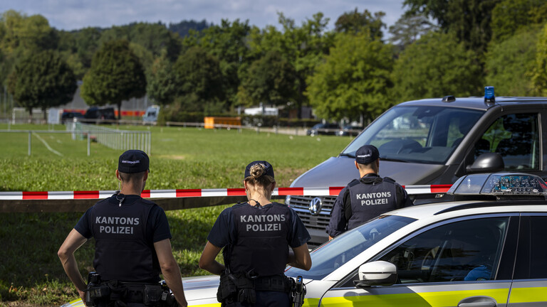 Drei Polizisten und zwei Polizeiwagen stehen vor der dem Absperrband an einer Pferderennbahn, auf der das Aarauer Rave stattgefunden hatte (Aarau, Schweiz, 30.08.2026)
