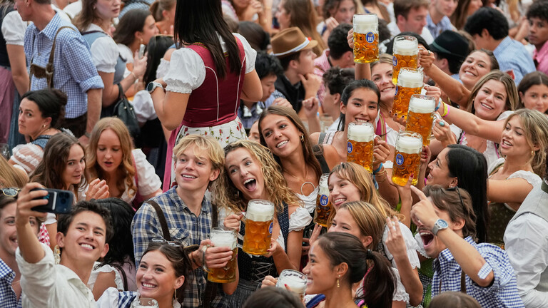 Bei der Eröffnung des Oktoberfests am 19. September 2026 sitzen und stehen junge Menschen an einem Tisch. Sie heben ihre Bierkrüge in die Höhe. Eine Person macht ein Selfie.