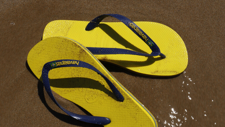 Foto von gelben Flipflops mit dunkelblauen Riemchen der Marke Havaianas am Strand, die von Wasser überspült werden