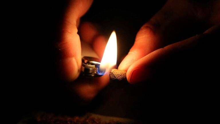 Zwei Hände im Dunkeln: Die eine Hand hält ein Stück Marihuana, die andere ein Feuerzeug mit Flamme.