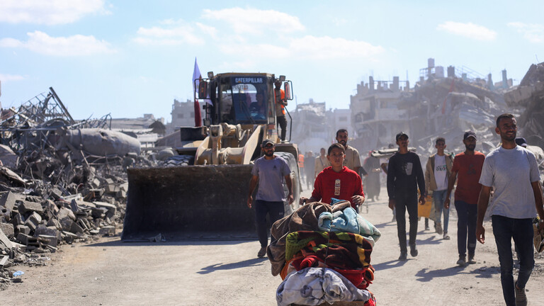 Menschen, die auf einem Karren ihr Hab und Gut transportieren, im Hintergrund ein Bulldozer, der Trümmer in Gaza-Stadt zur Seite räumt.