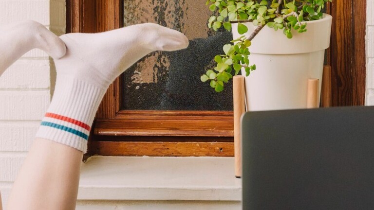 Gelangweilte Frau mit hochgelegten Füßen am Laptop am Tisch im Homeoffice.