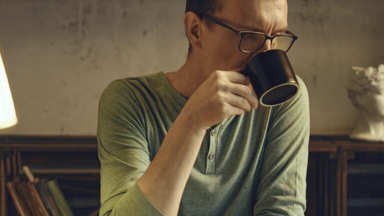 Ein Mann mittleren Alters sitzt an einem Schreibtisch in einem gemütlichen Zimmer und schlürft einen Kräutertee aus einer Tasse. Das sanfte Licht einer Lampe beleuchtet das Buch, das er vor dem Schlafengehen liest