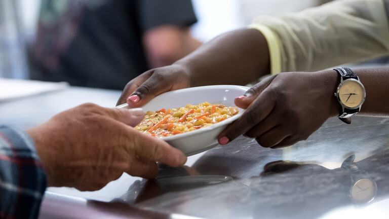Foto der Hände einer Frau, die eine gefüllte Suppenschüssel reicht, die von der Hand eines alten Mannes entgegengenommen wird