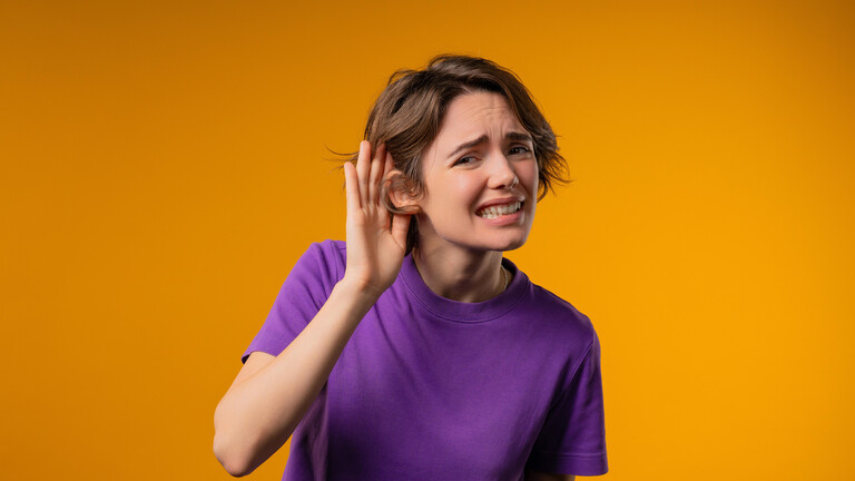 Foto einer jungen Frau im lila T-Shirt vor einem orangenen Hintergrund, die die Hand ans Ohr legt und das Gesicht verzieht, als ob sie etwas schlecht hören kann