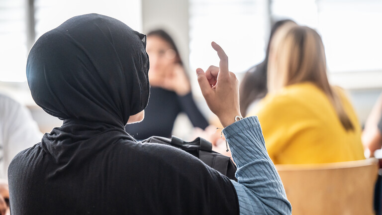 Mädchen mit Hijab (Kopftuch) in einem Klassenraum, Ansicht von hinten, sie hebt den Finger