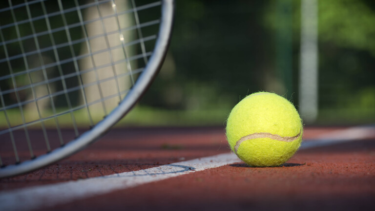 Nahaufnahme eines Tennisballs auf einem roten Sandplatz. Der Tennisball liegt auf der rechten Seite des Platzes,