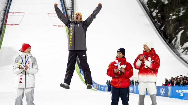 Philipp Raimund hat Gold beim Skispringen gewonnen. Der Sportler steht bei der Siegerehrung auf dem Treppchen und springt hoch in die Luft.