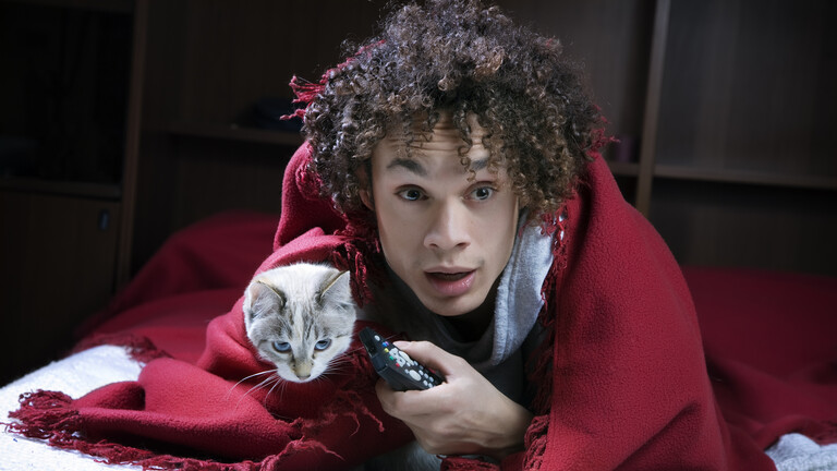 Mann mit Katze unter roter Kuscheldecke und Fernbedienung in der Hand schaut Horrorfilm und schaut verängstigt