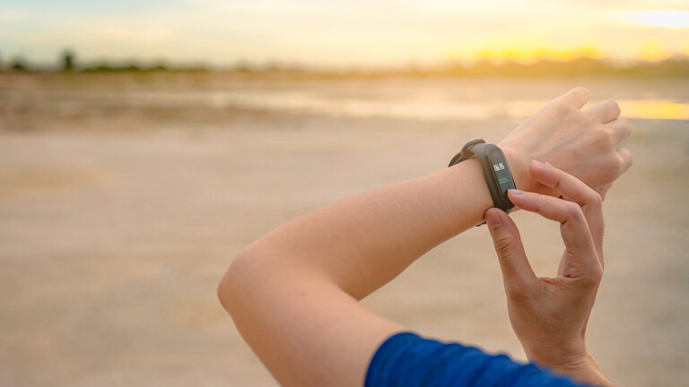 Foto der Arme einer Frau in einem blauen Tshirt, die mit der rechten Hand Einstellungen an einem Fitnesstracker vornimmt, den sie am linken Handgelenk trägt, im Hintergrund unscharf ein Strand