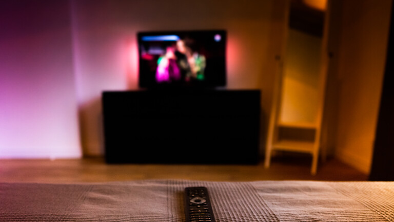 Eine Fernbedienung liegt auf einem Bett, der Fernseher ist an