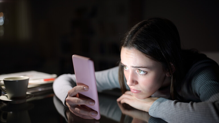 Eine junge Frau schaut auf ein Smartphone; sie stützt sich auf einem Tisch ab und hat den Kopf auf eine Hand gelegt.