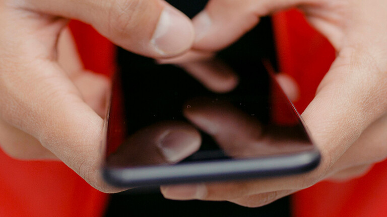 Foto der Hände eines jungen Mannes, der eine rote Daunenjacke trägt und mit beiden Daumen auf einem Smartphone tippt