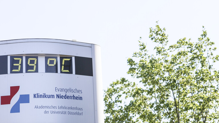 Thermometer vor Krankenhaus zeigt 39 Grad, daneben ein Baum