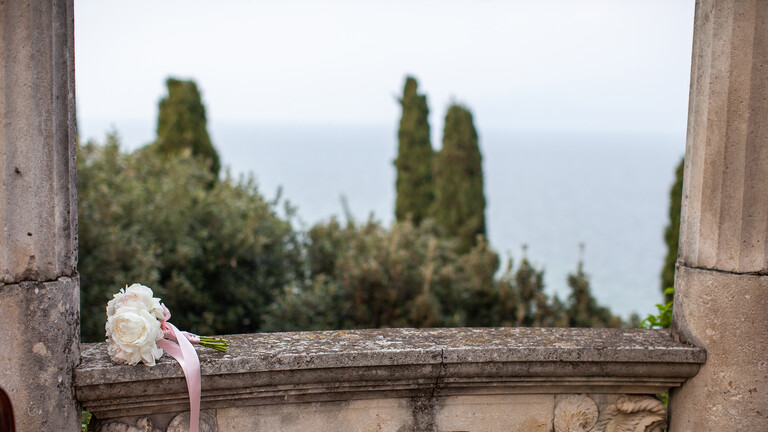 Ein Hochzeitsstrauß liegt auf dem Geländer eines antiken Gebäudes in Italien. Dahinter offenbart sich ein Blick aufs Meer.