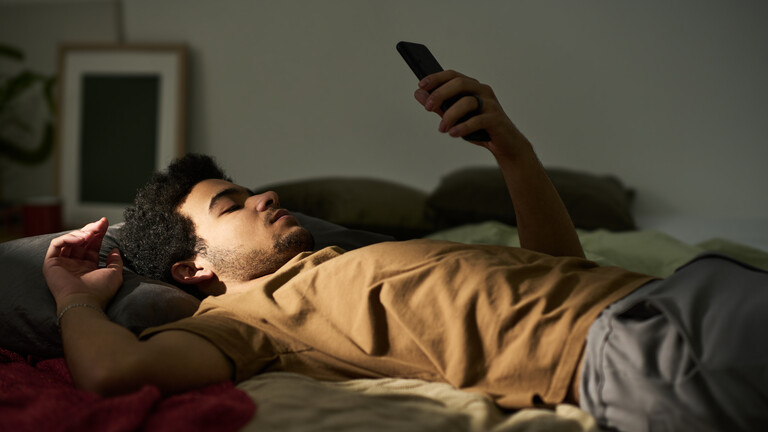 Ein Mann liegt auf dem Rücken im Bett. Sein linker Arm ist aufgestützt und er hält ein Smartphone in die Höhe, auf das er blickt. Es wirkt ruhig und etwas traurig.