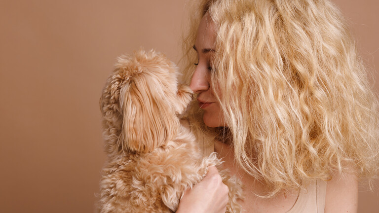 Eine Frau und ihr Hund schauen sich an, die Nase berühren sich. Beide haben gelocktes Haar beziehungsweise Fell und ein ähnliche Farbe, ein Blond-Braun. Die Frau wirkt glücklich.