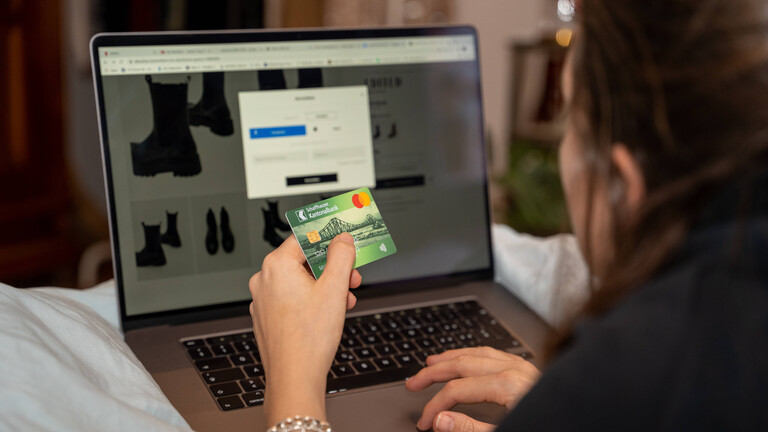 Eine Frau sitzt vor einem Laptop und hat eine Shoppingseite geöffnet. in der linken Hand hält sie ihre Kreditkarte, während sie mit der rechten tippt.
