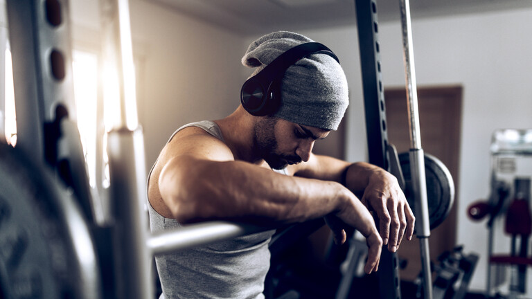 Mann mit Mütze und Kopfhörer im Fitnesstudio, Pause an der Hantel