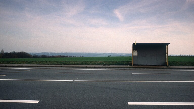 Eine leere Bushaltestelle, davor eine leere Straße, dahinter flache Landschaft. Das Bild strahlt Stillstand aus.