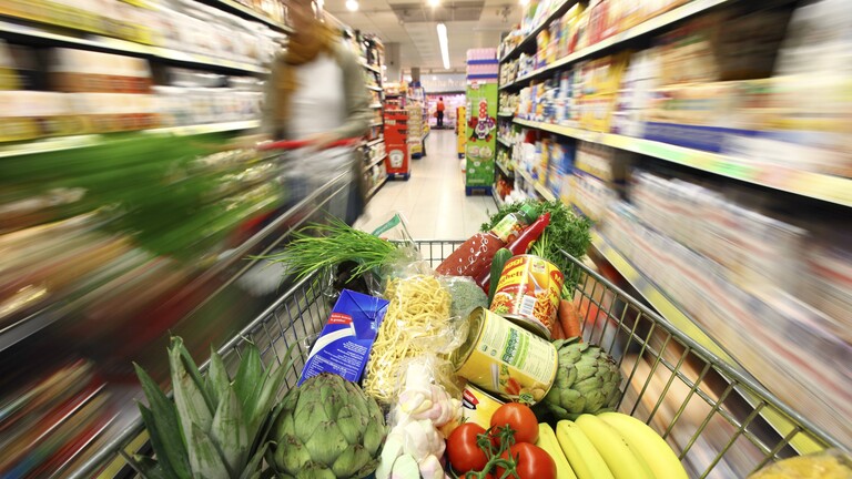 In einem Supermarkt wird ein voller Einkaufswagen durch einen Gang mit diversen Lebensmittelprodukten geschoben. Der Hintergrund ist verschwommen; es wirkt als ob der Einkaufswagen schnell bewegt wird.