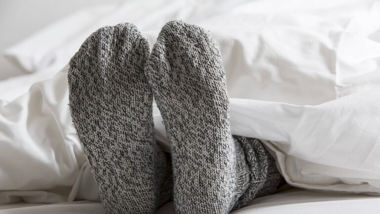 Unter einer weißen Bettdecke schauen Füße hervor, die in warmen grauen Wollsocken stecken.