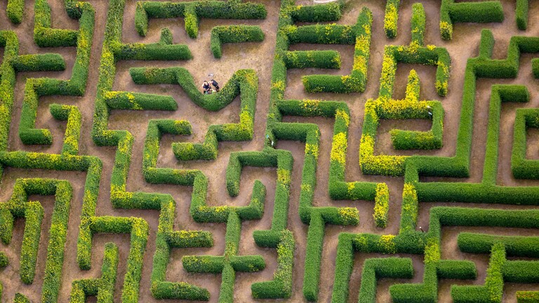 Aus der Vogelperspektive Blick auf ein Labyrinth aus Hecken. Zwei Menschen stehen in einem der Gänge.