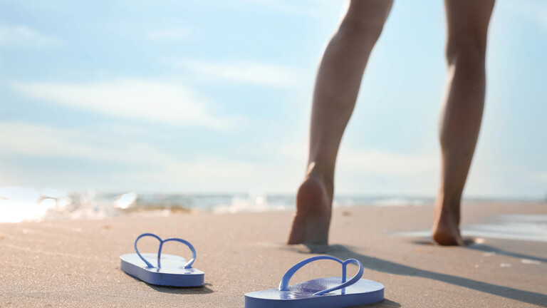 Frau läuft barfuß am Strand, links der Ozean, vorn im Bild ihre Flip-Flops