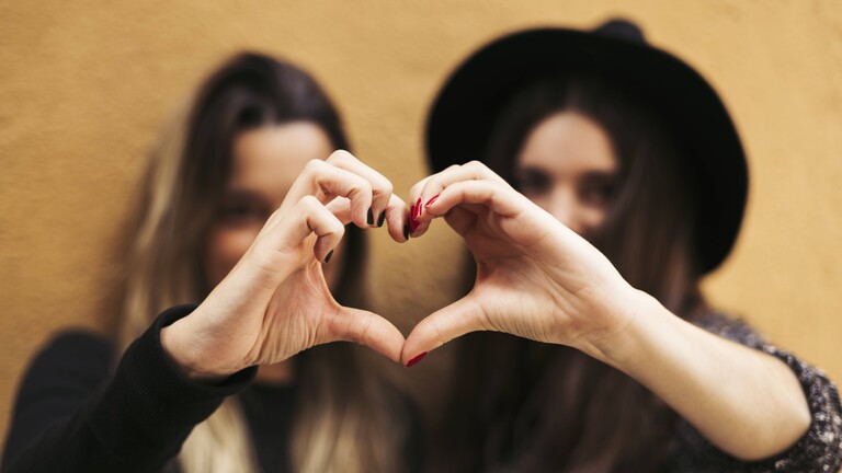 Zwei Frauen stehen nebeneinander. Beide strecken ihre rechte beziehungsweise linke Hand nach vorne. Die beiden Hände bilden ein Herz.