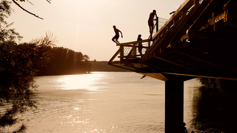 Menschen springen in der Abendsonne von einem Steg am Flughafensee in Berlin.