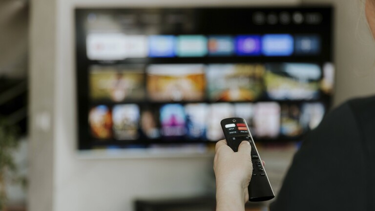 Foto einer Hand mit einer Fernbedienung vor dem unscharfen Hintergrund eines Smart-TVs mit Auswahlkacheln für Medieninhalte