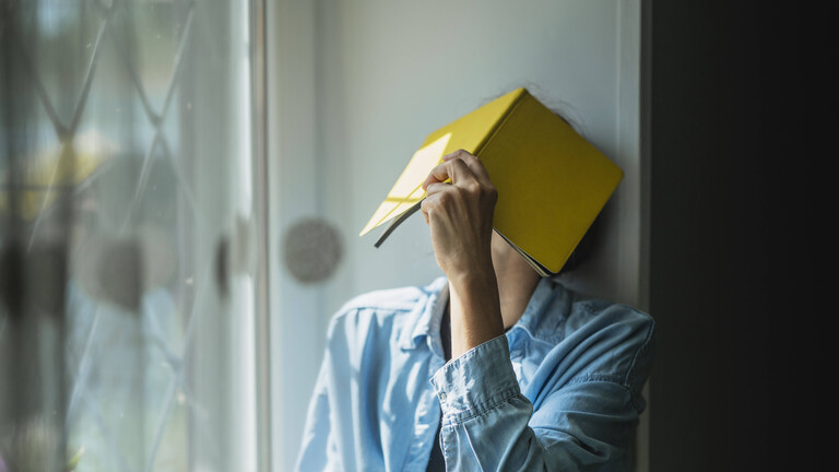 Eine Frau sitzt am Fenster und hält sich ein Buch vors Gesicht.