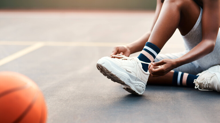 Nahaufnahme der Füße einer jungen Frau, die auf einem Hallenboden sitzt und ihre Schnürsenkel bindet, im Vordergrund liegt unscharf ein Basketball