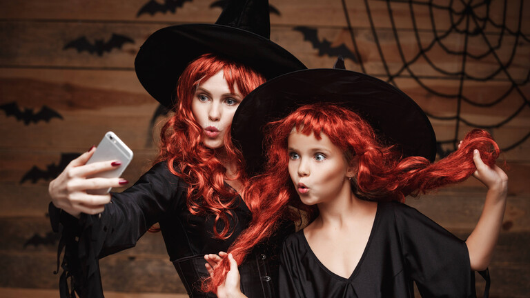 Mutter und ihre Tochter mit langen roten Haaren in Hexenkostümen machen ein Selfie mit dem Smartphone, bereit für die Halloween-Feier.