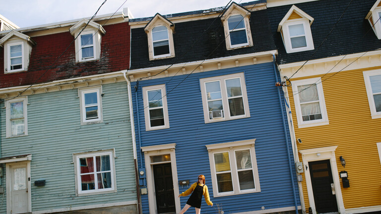 Eine Frau in einem langen gelben Pullover schreitet fröhlich über eine Straße, die von farbenfrohen Reihenhäusern gesäumt ist. Die Kulisse unterstreicht den historischen Charme von St. John’s, Neufundland und Labrador, Kanada