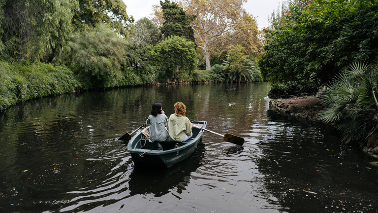 Lesbisches Pärchen in einem Ruderboot auf einem Weiher in Barcelona. Ringsum viel Grün.
