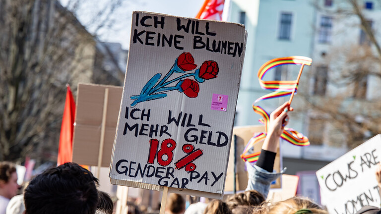 Frauen bei einer Demonstration zum Internationalen Frauentag in Berlin halten ein Plakat in den Händen. Darauf steht: Ich will keine Blumen, ich will mehr Geld. 18 Prozent Gender Pay Gap.