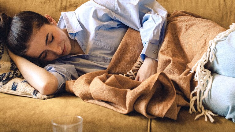 Eine Frau liegt mit geschlossenen Augen seitlich auf dem Sofa, hat die Beine angezogen und deckt sich mit einer Decke zu.