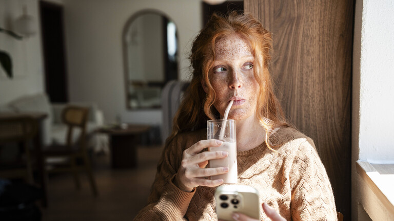 Eine junge Frau sitz am Fenster und blickt nach draußen. In der einen Hand hält sie ein Smartphone, in der anderen ein Glas mit Milch. Sie trinkt daraus mit einem Strohhalm.