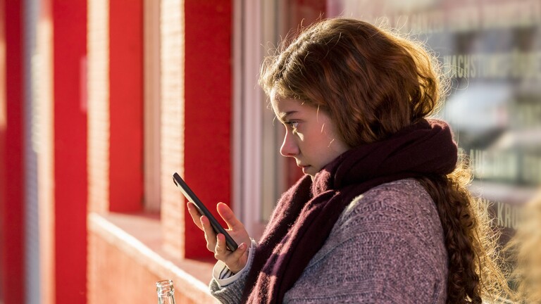 Eine Jugendliche steht auf einer roten Brücke. Sie hat in der einen Hand ein Handy, in der anderen eine Limonade.