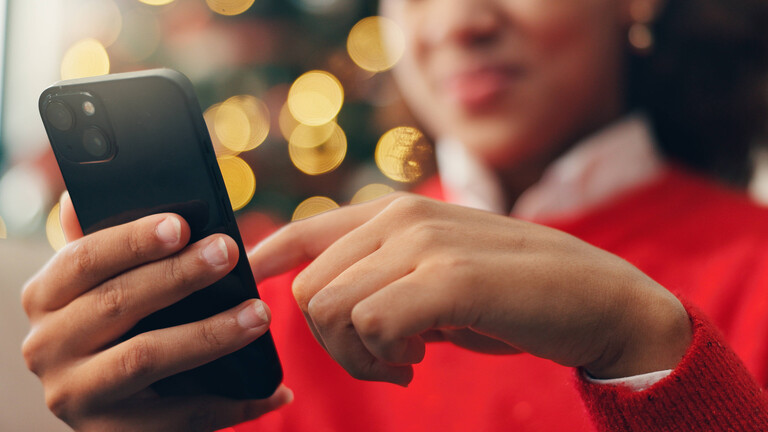 IMAGO / YAY ImagesNahaufnahme der Hände einer Frau mit rotem Pulli, die vor einem unscharfen Weihnachtsbaum auf einemSmartphone tippt
