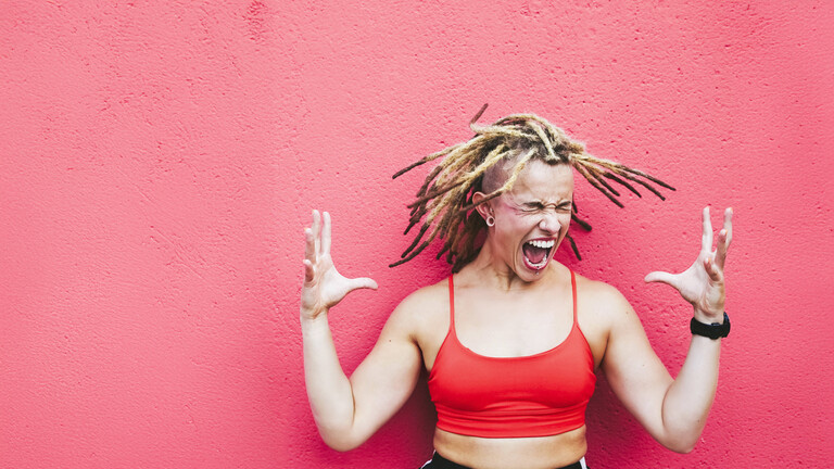Foto einer jungen, sportlichen Frau in Sportkleidung, die in einer hockenden Kraftpose die Hände hebt, den Kopf schüttelt und schreit