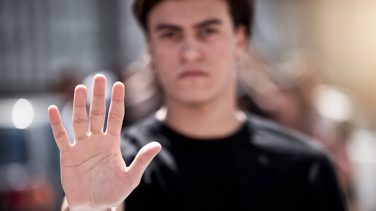 Foto eines jungen Mannes, der die Hand hebt und so "Stopp" signalisiert - Die Hand vorne ist scharf, der Mann dahinter unscharf.