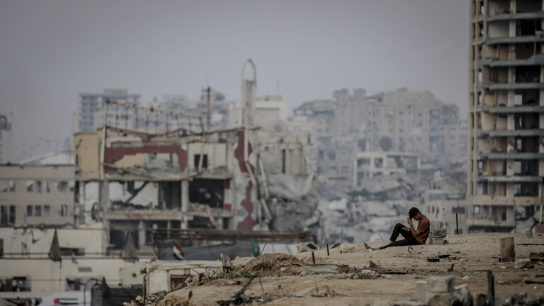 In Gaza Stadt sitzt ein junger Mann auf dem Boden, er hat die Beine angezogen und den Kopf auf seine Hände gestützt. Im Hintergrund sind zerstörte Gebäude zu sehen.
